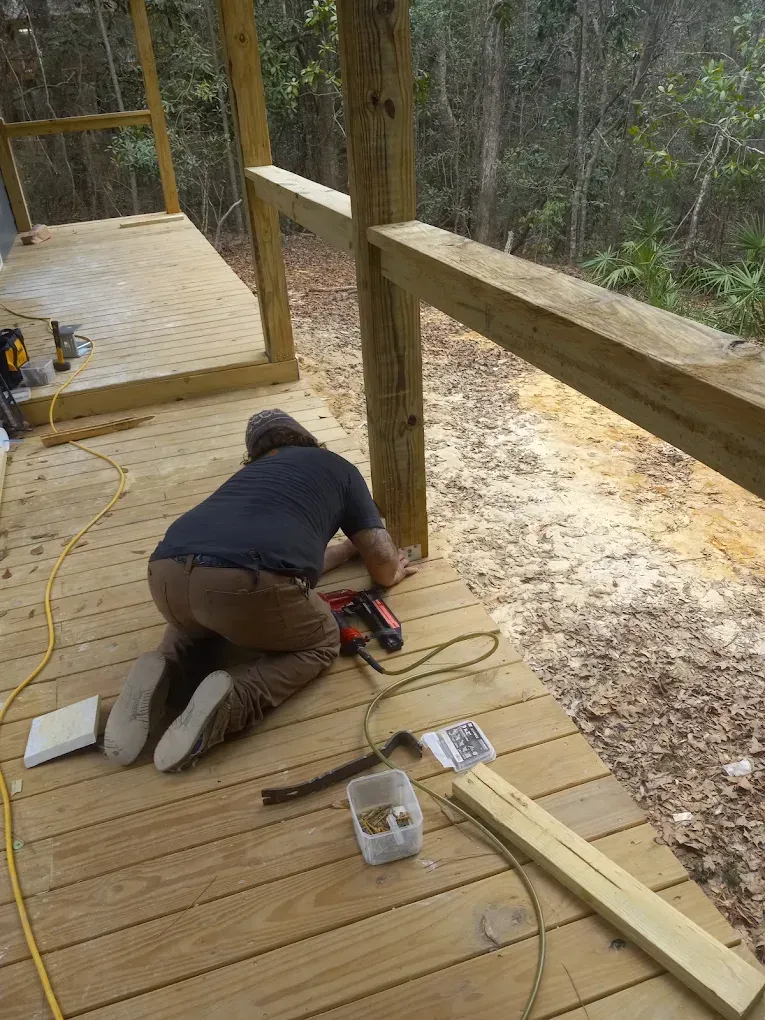 Person kneeling on a wooden deck using a red power tool beside a railing in a wooded area