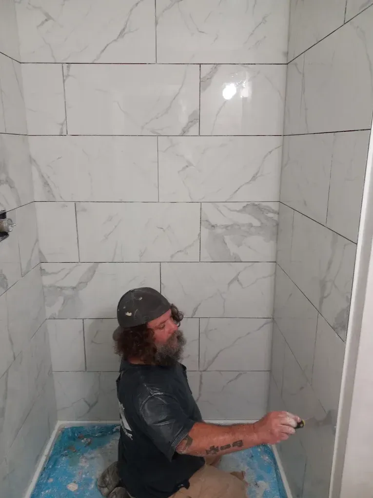 Person kneeling in a tiled shower stall, wearing a dark shirt and cap, holding a tool while working on the wall.