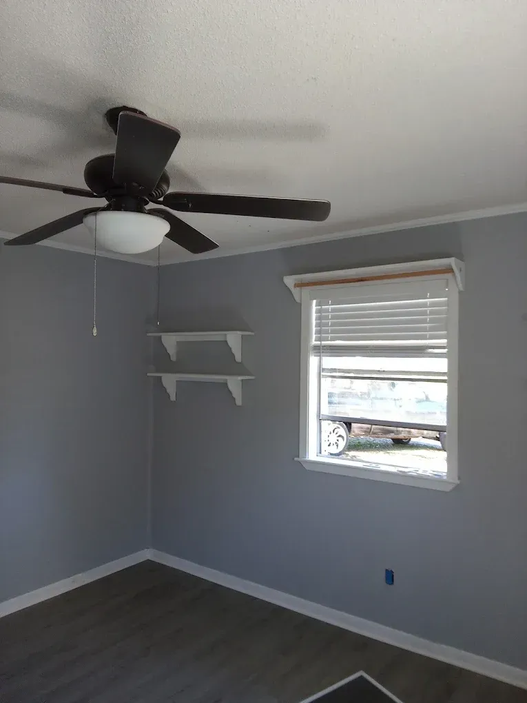 Empty gray bedroom with a ceiling fan, white shelves, and a window with blinds.