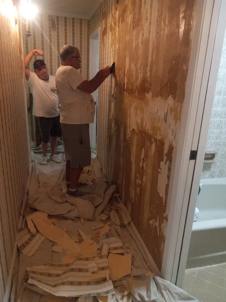 Two people stripping wallpaper in a narrow hallway with debris on the floor.