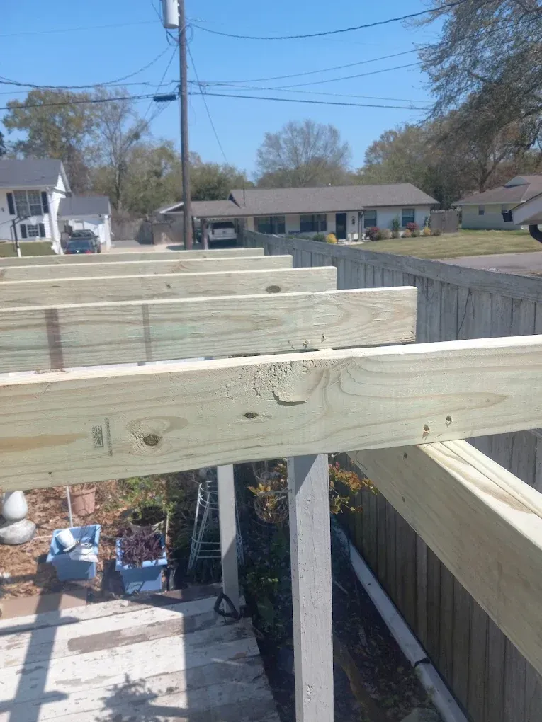 Wooden deck framing under construction in a backyard, with houses and blue sky in the background.