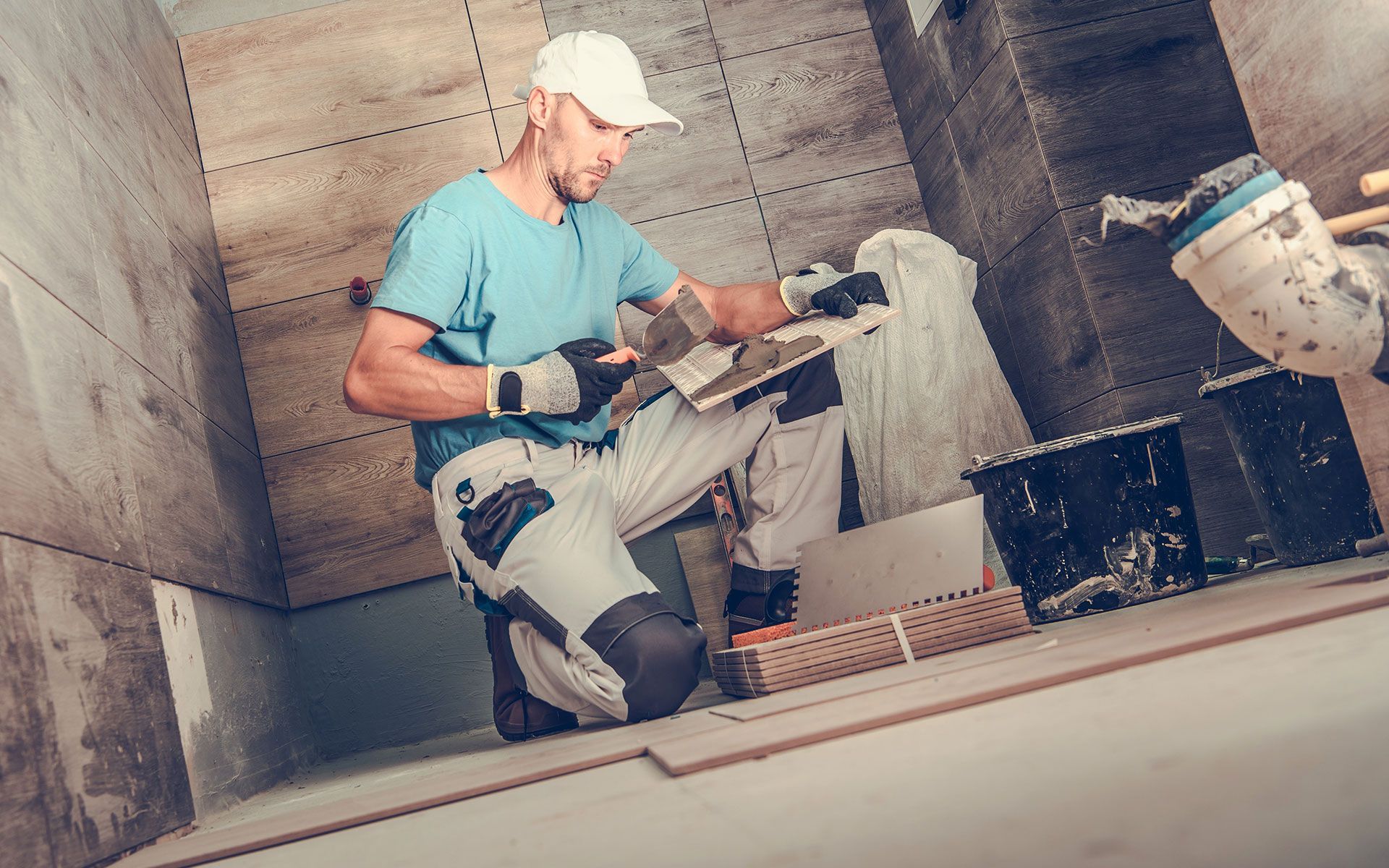 Man tiling a wall, kneeling, using a trowel. Inside a room, wearing a cap and work clothes.