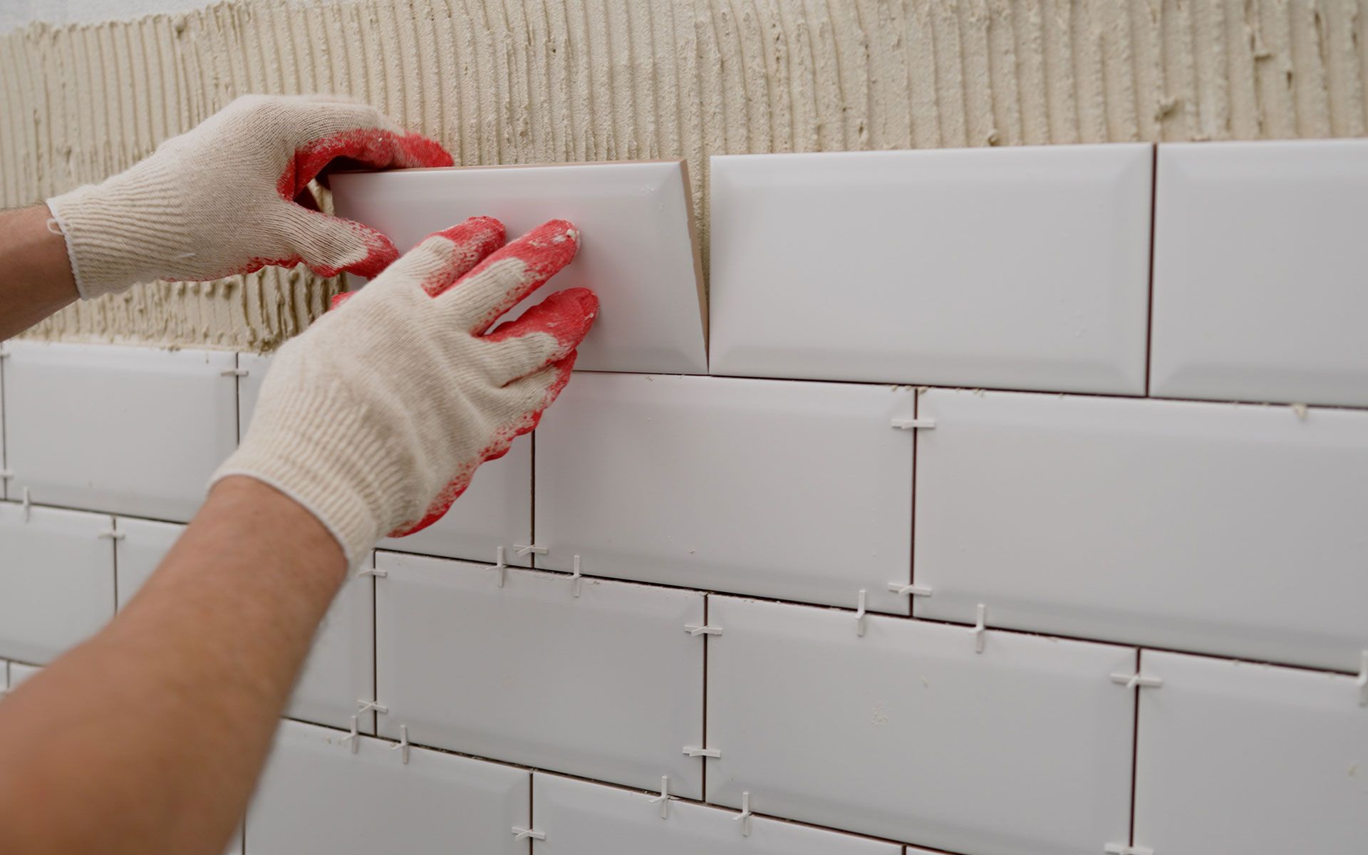 Hands in gloves placing white subway tile on a wall, using tile spacers.
