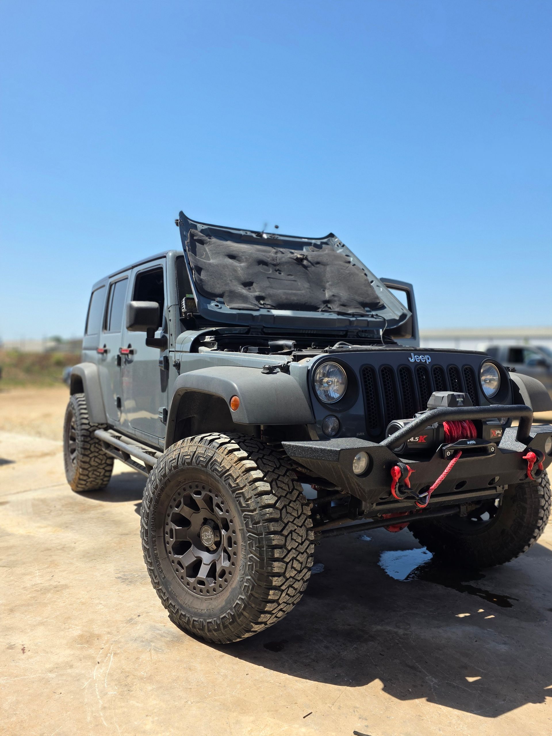 Black Jeep Wrangler with raised hood and off-road tires parked on a dirt road under a clear blue sky