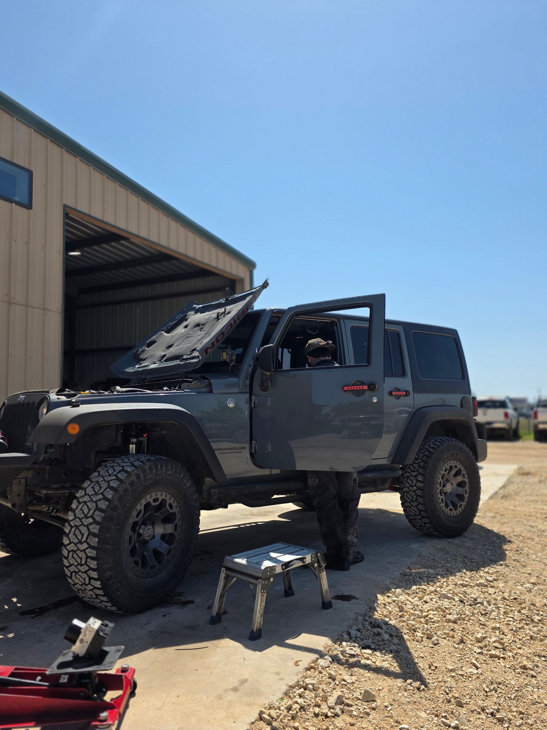 Gray off-road Jeep with open hood and driver door, person standing beside it outside a workshop