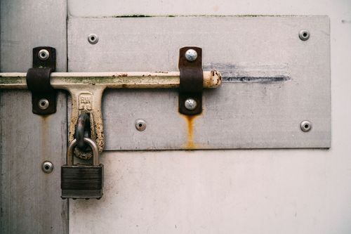 Weathered metal door latch secured with a padlock on a white door panel.