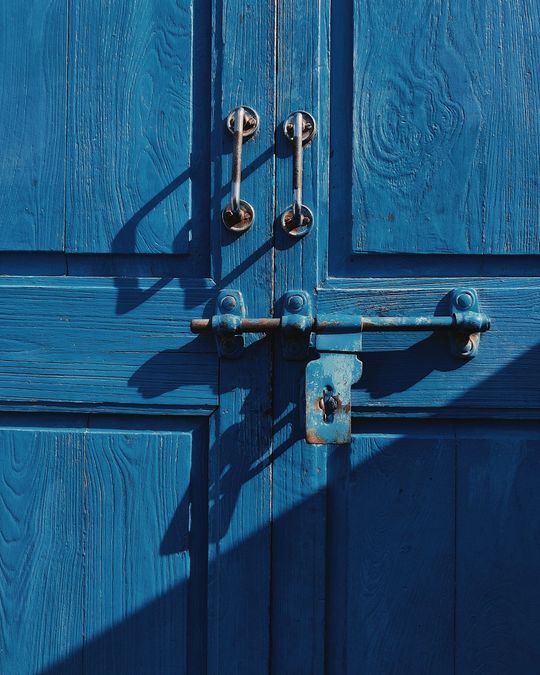 Blue wooden doors with metal latches and a padlock, lit by sharp sunlight and shadow.
