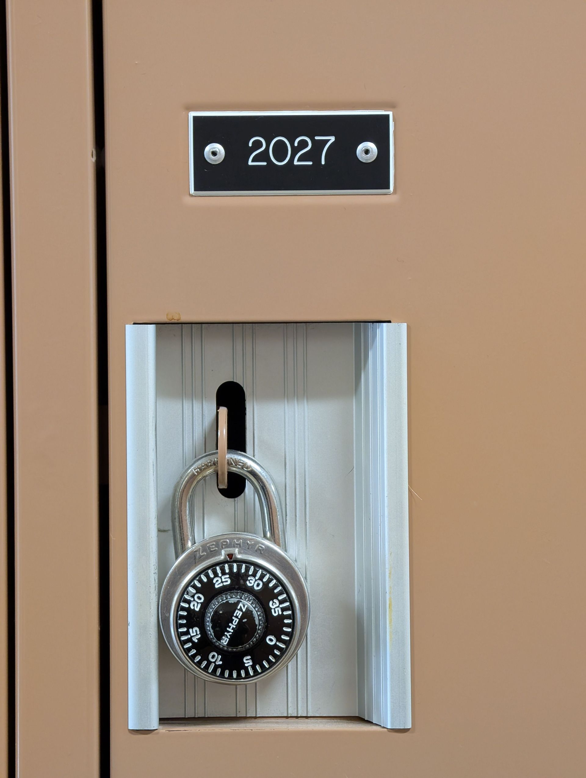 Combination padlock hanging in a locker labeled 2027, with a white recessed handle area.