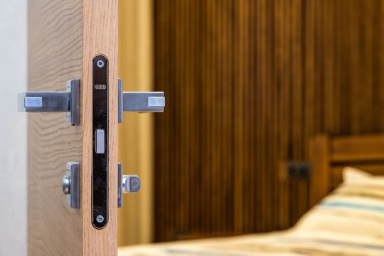 Close-up of a modern door latch on a partially open wooden door, with a blurred bedroom in the background.