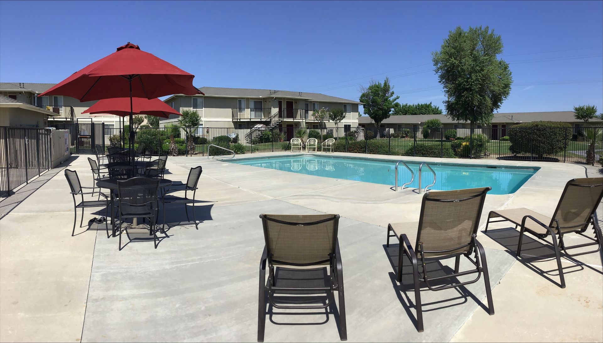 Poolside scene with chairs, tables, red umbrellas, and apartment buildings under a clear blue sky.