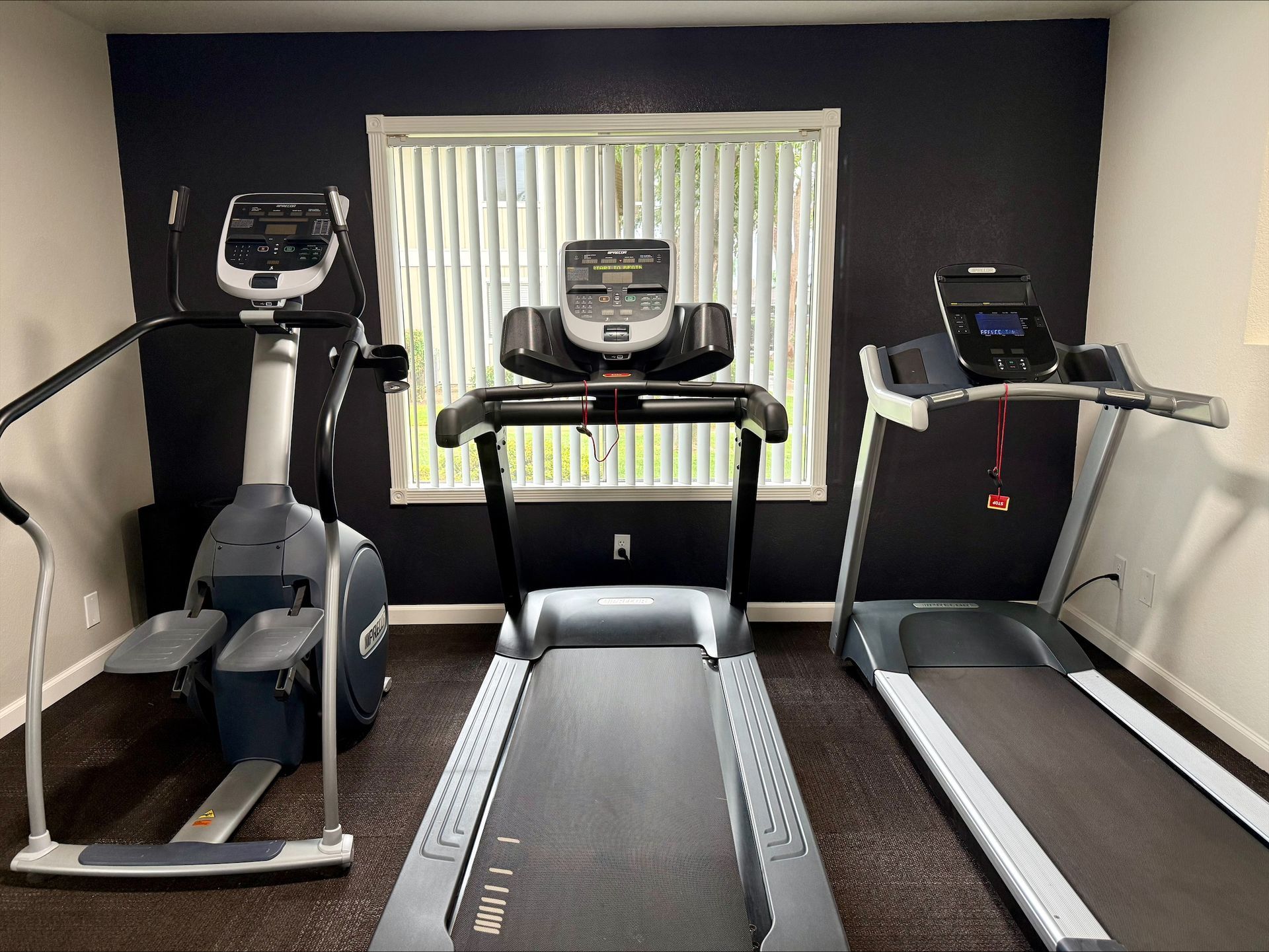 Gym with an elliptical machine, treadmill, and stationary bike in front of a window with blinds. Dark wall.