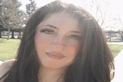 Close-up of a woman outdoors in sunlight, with trees and grass in the background.