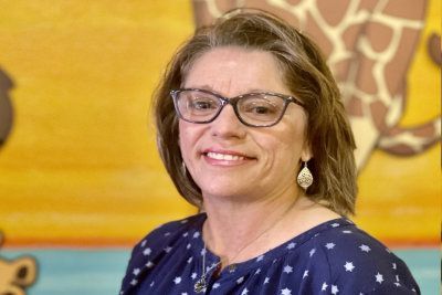 Smiling woman wearing glasses in a blue patterned top, seated against a warm-toned mural background