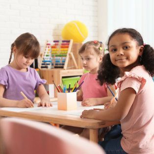 Children drawing at a table in a bright classroom with colorful art supplies.