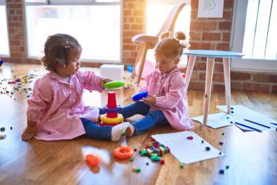 Two children in pink smocks play with colorful toys on the floor of a bright classroom.
