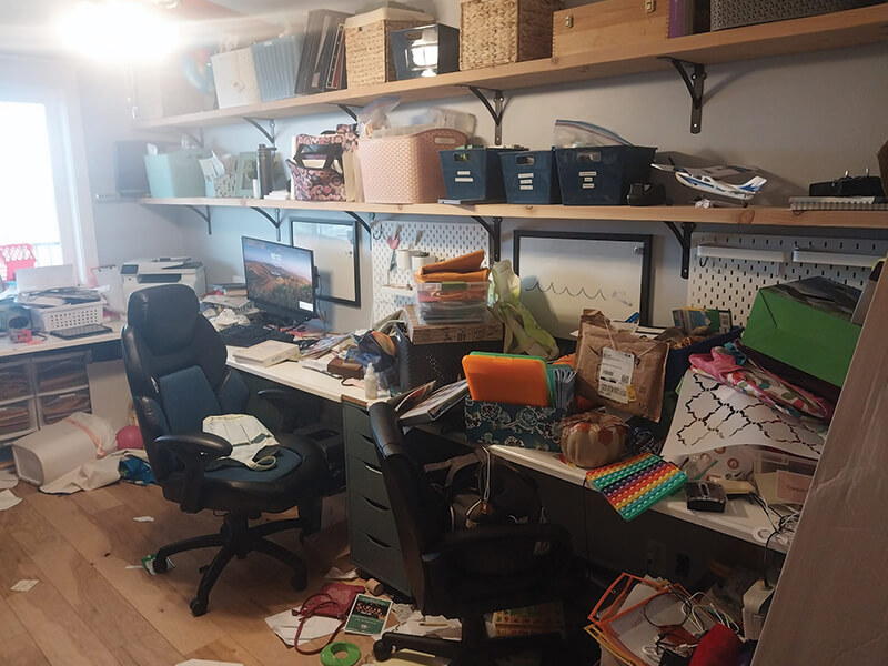 Cluttered home office with desk, chair, shelves, and various containers. Overwhelmed with items.