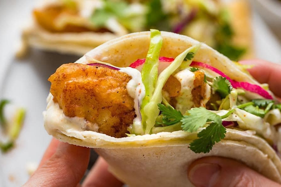 Close-up of a hand holding a fish taco filled with breaded fish, slaw, cilantro, and sauce.