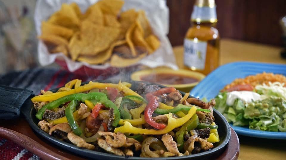 Sizzling fajitas with colorful peppers, tortilla chips, salsa, rice, salad, and a beer bottle on a table.