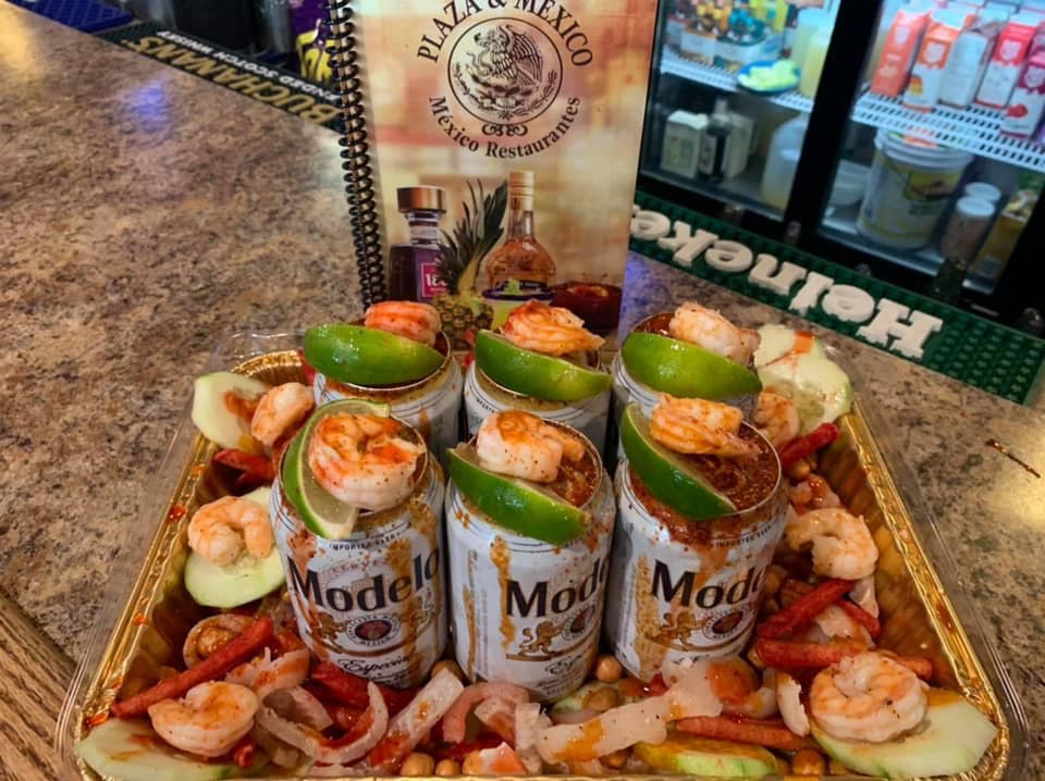 Tray of Modelo beer cans with shrimp, lime, and garnishes. Plaza & Mexico menu in background.