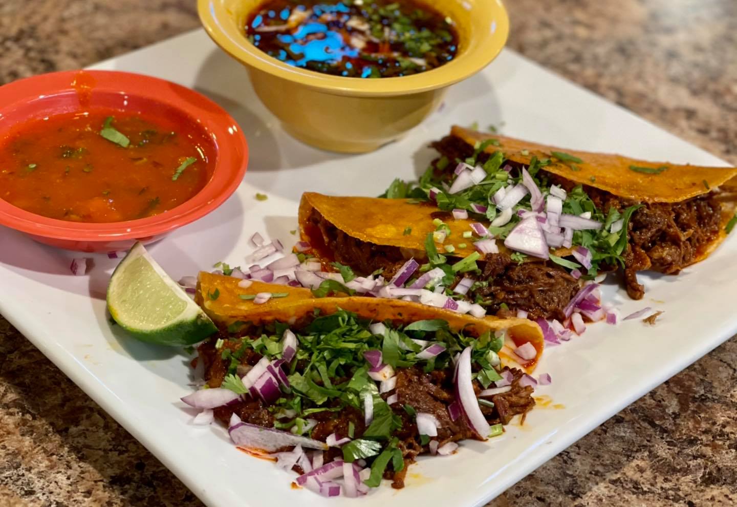 Birria tacos with consommé and lime wedge on a white plate.