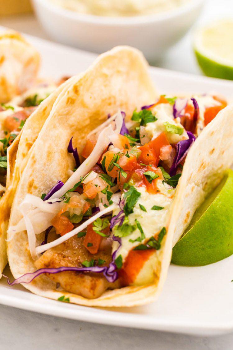 Fish tacos with toppings, lime, and sauce on a white plate.