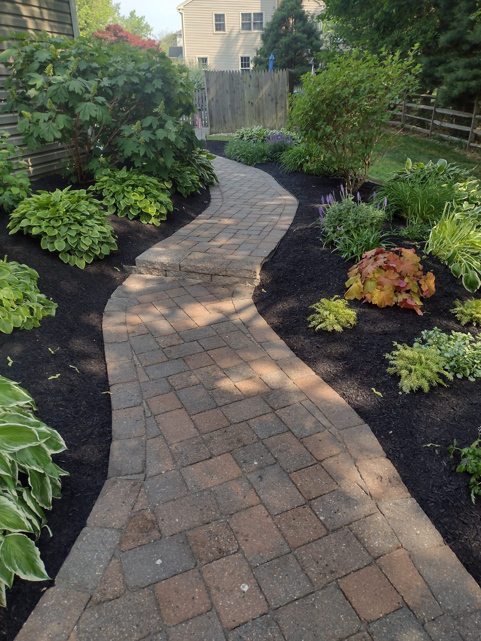 Curved brick garden path bordered by dark mulch and green shrubs, leading to a wooden fence.