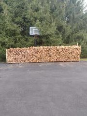 Stack of firewood along an asphalt driveway with a basketball hoop and trees in the background