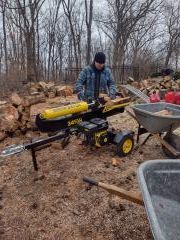 Person operating a yellow log splitter beside a wheelbarrow in a wooded outdoor area