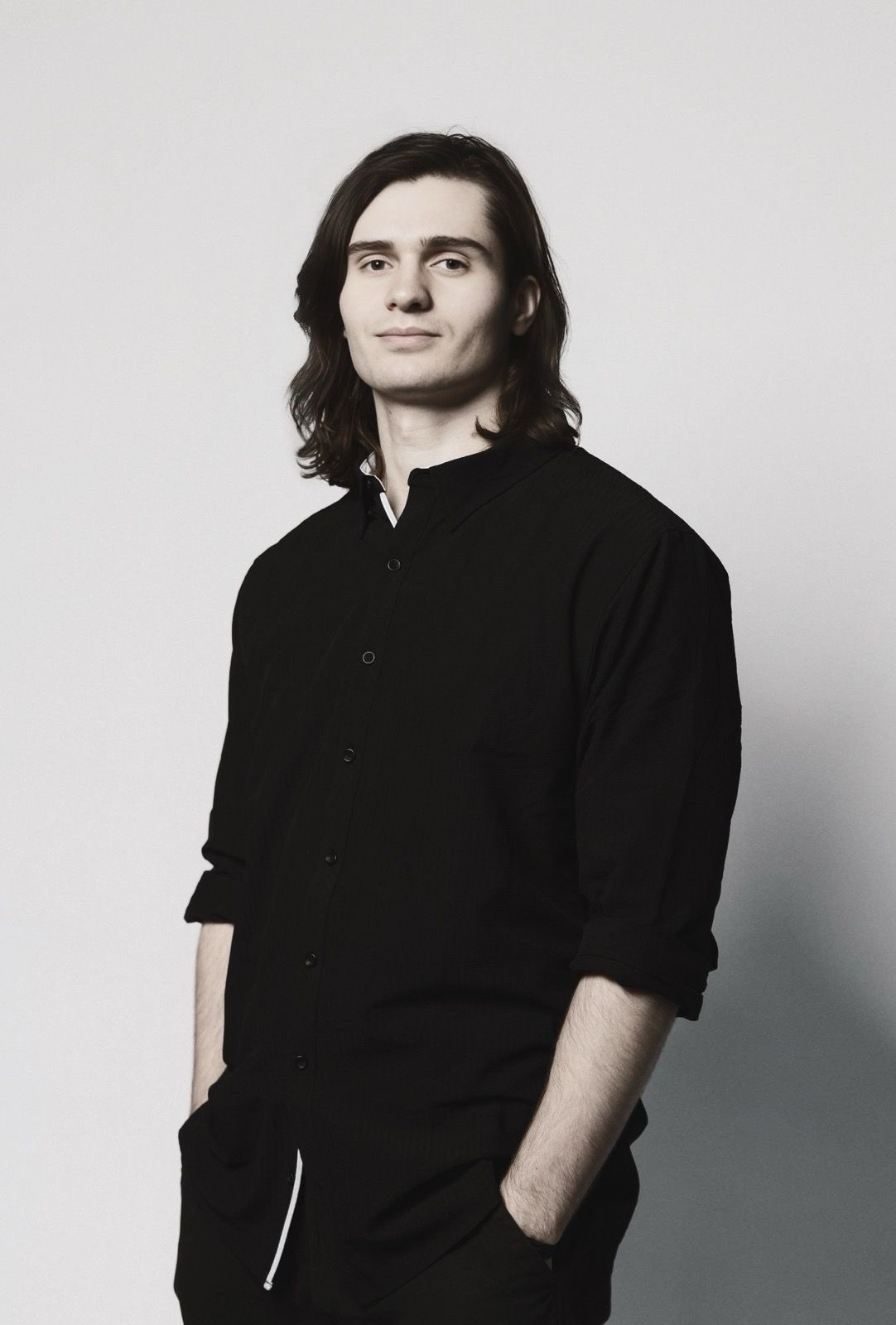 A person with shoulder-length dark hair wearing a black shirt, standing against a plain, light background.