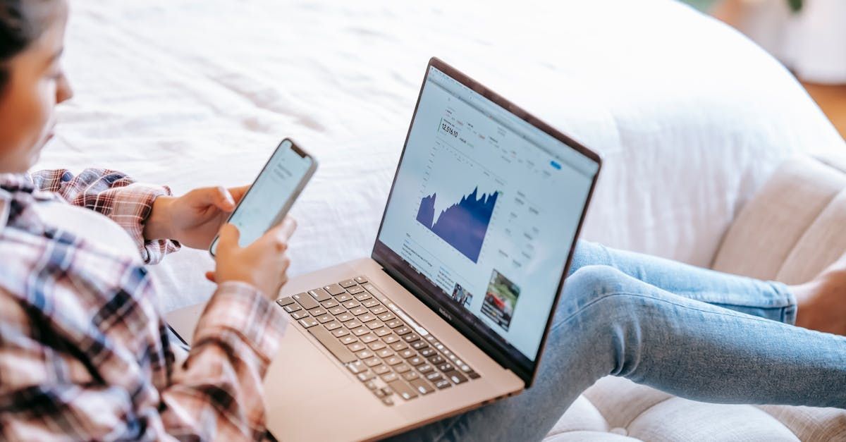 A woman is sitting on a couch using a laptop and a cell phone.