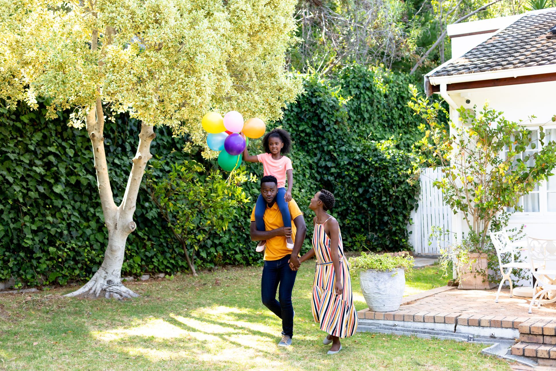 Three people walking in a sunny yard, one holding colorful balloons near a house