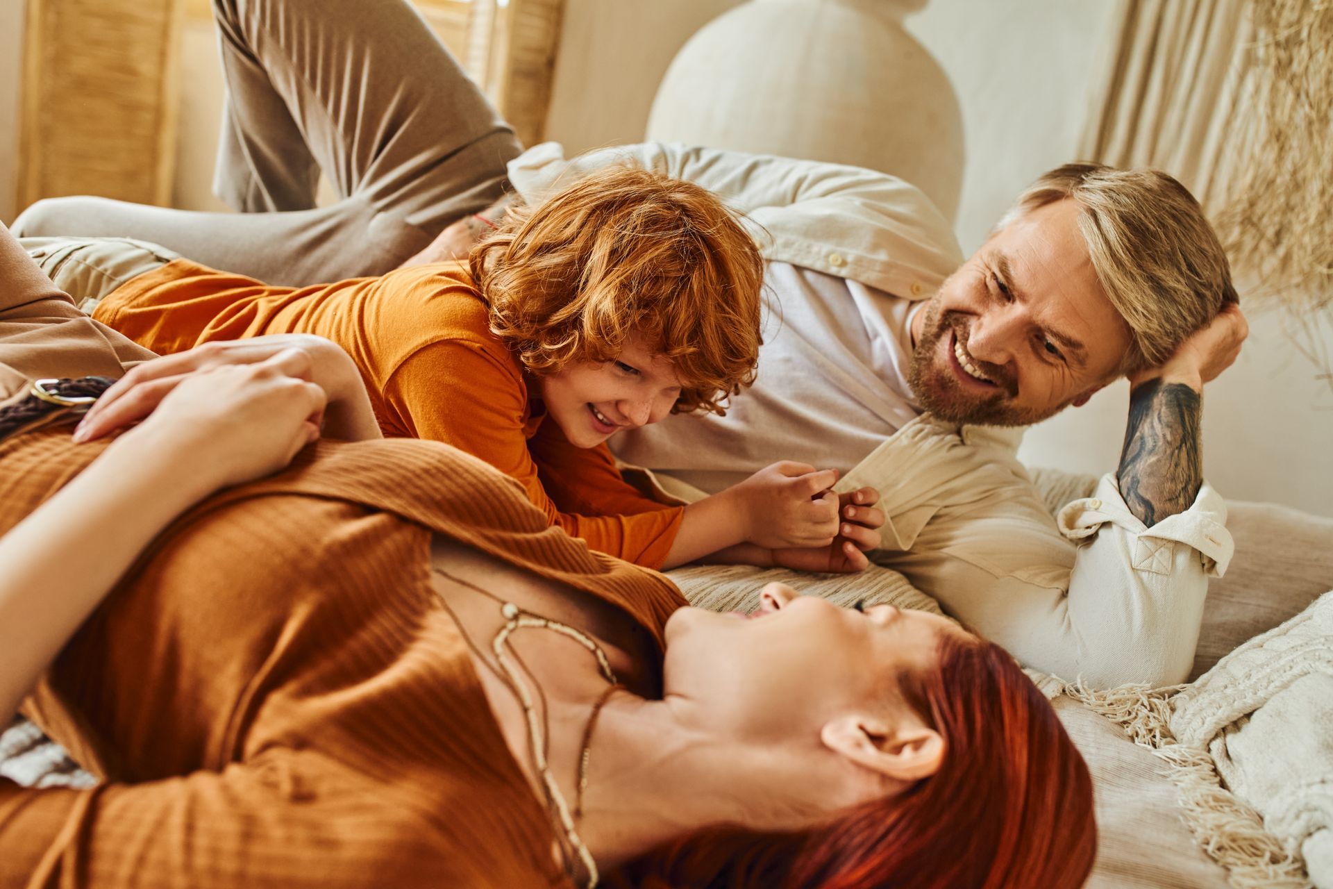 Three people relax on a couch, smiling and laughing together under a warm blanket.