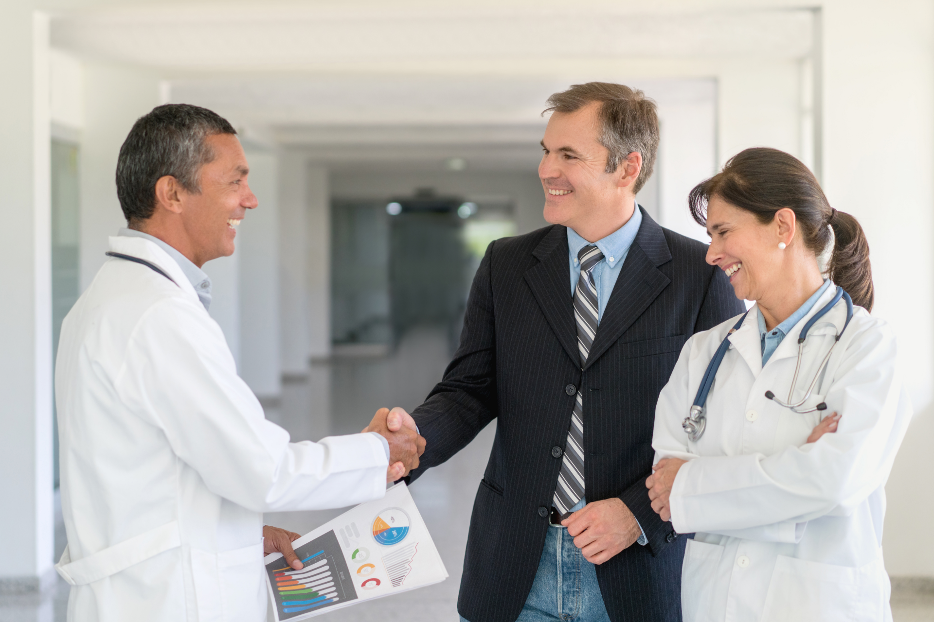 Doctor and businessman shaking hands in a bright hospital hallway, with a female doctor smiling nearby