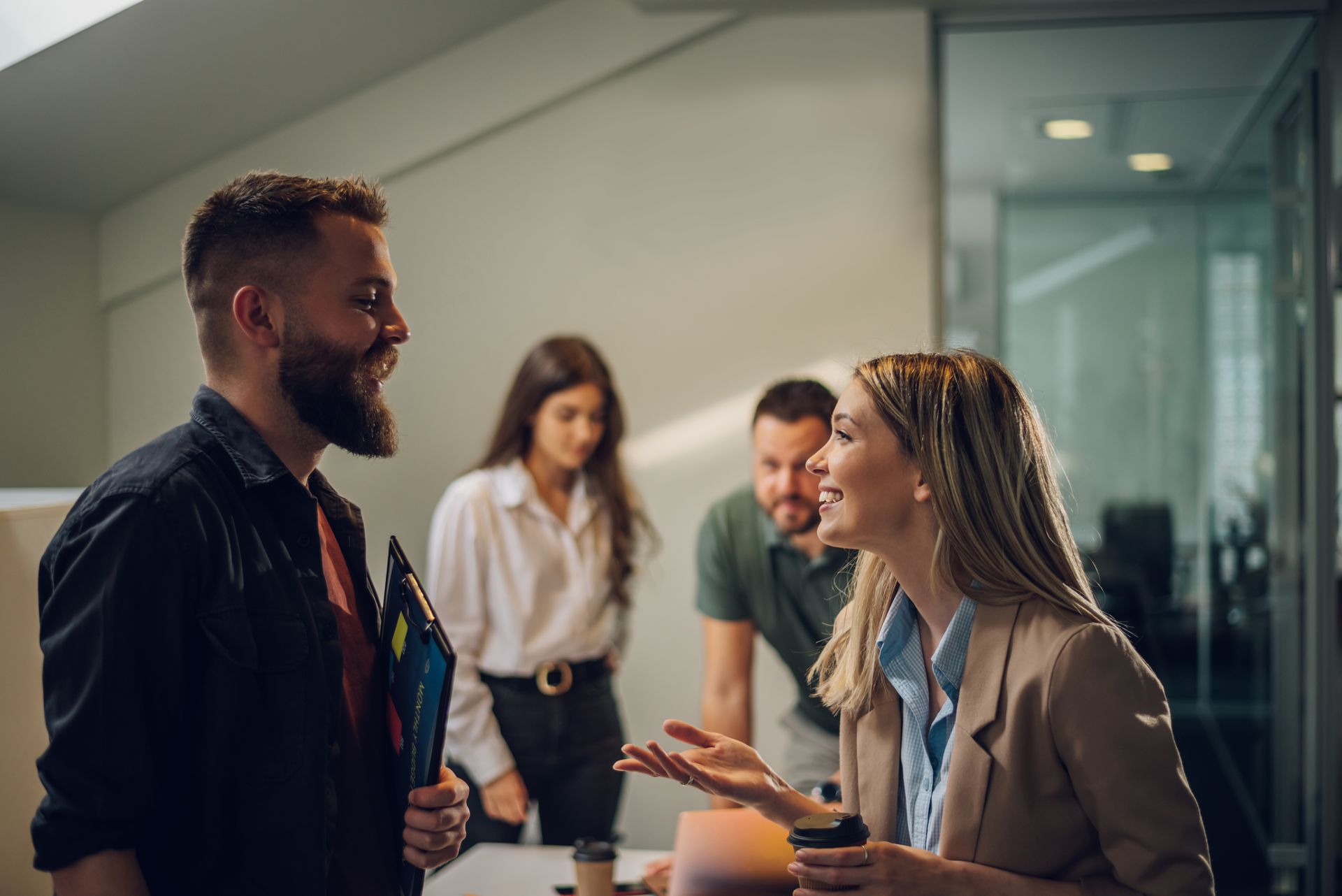 Two coworkers smiling and talking in a modern office, with others blurred in the background.