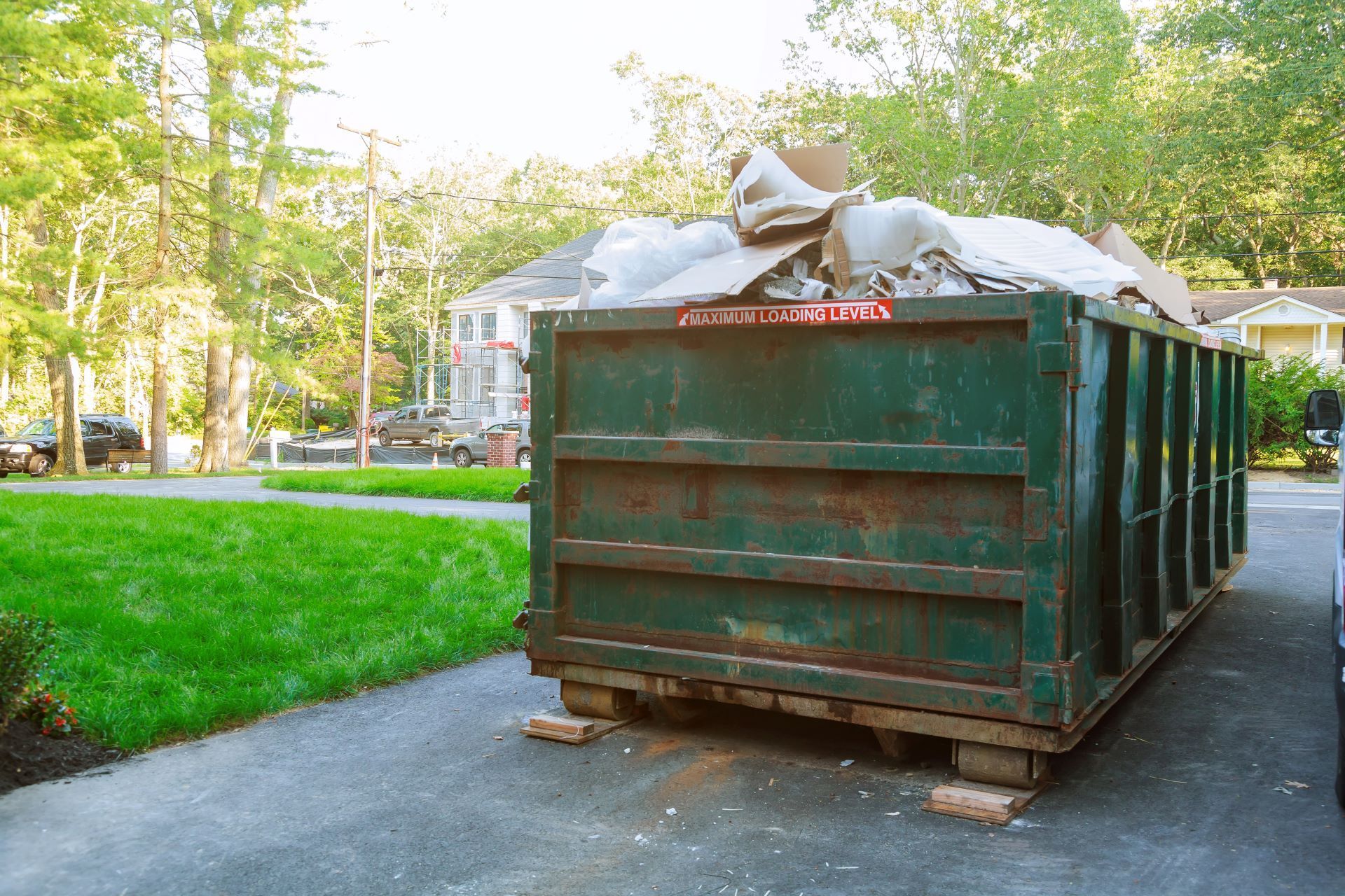 green dumpster in driveway filled to capacity. Trees, cars and a house in background.