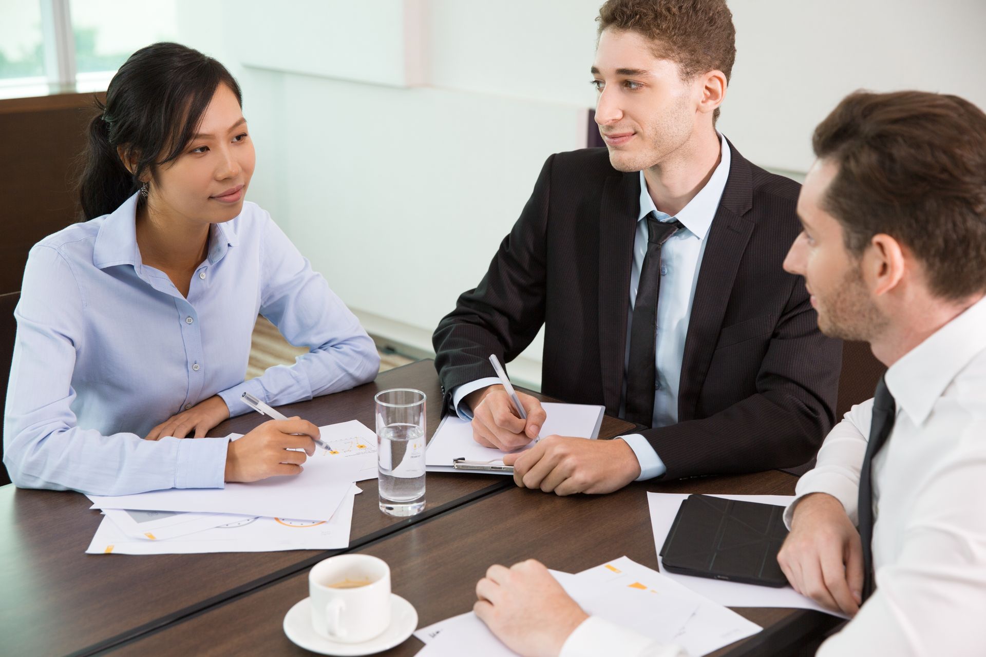 Three colleagues in a meeting around a table, discussing documents with coffee cups in a bright office.