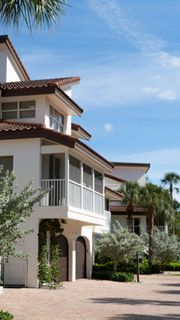 Mediterranean-style stucco house with balconies, arches, and palm trees along a gravel driveway