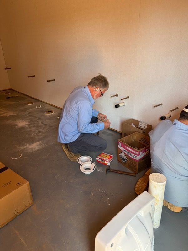 Person kneeling on a concrete floor, working with tools and supplies beside a wall in a bare room.