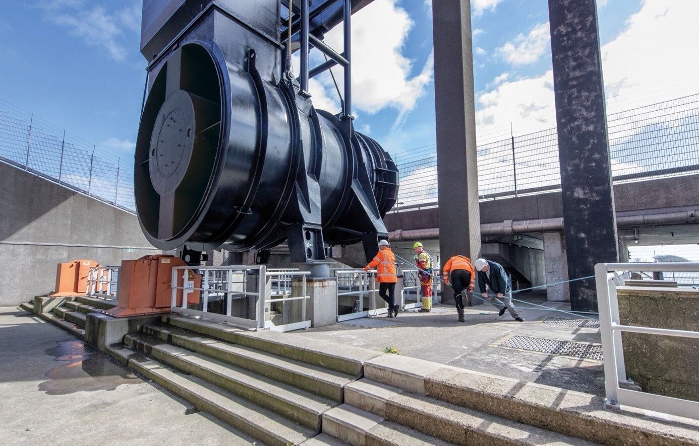 Een grote zwarte sluis met arbeiders in oranje hesjes, betonnen trappen en een brug.