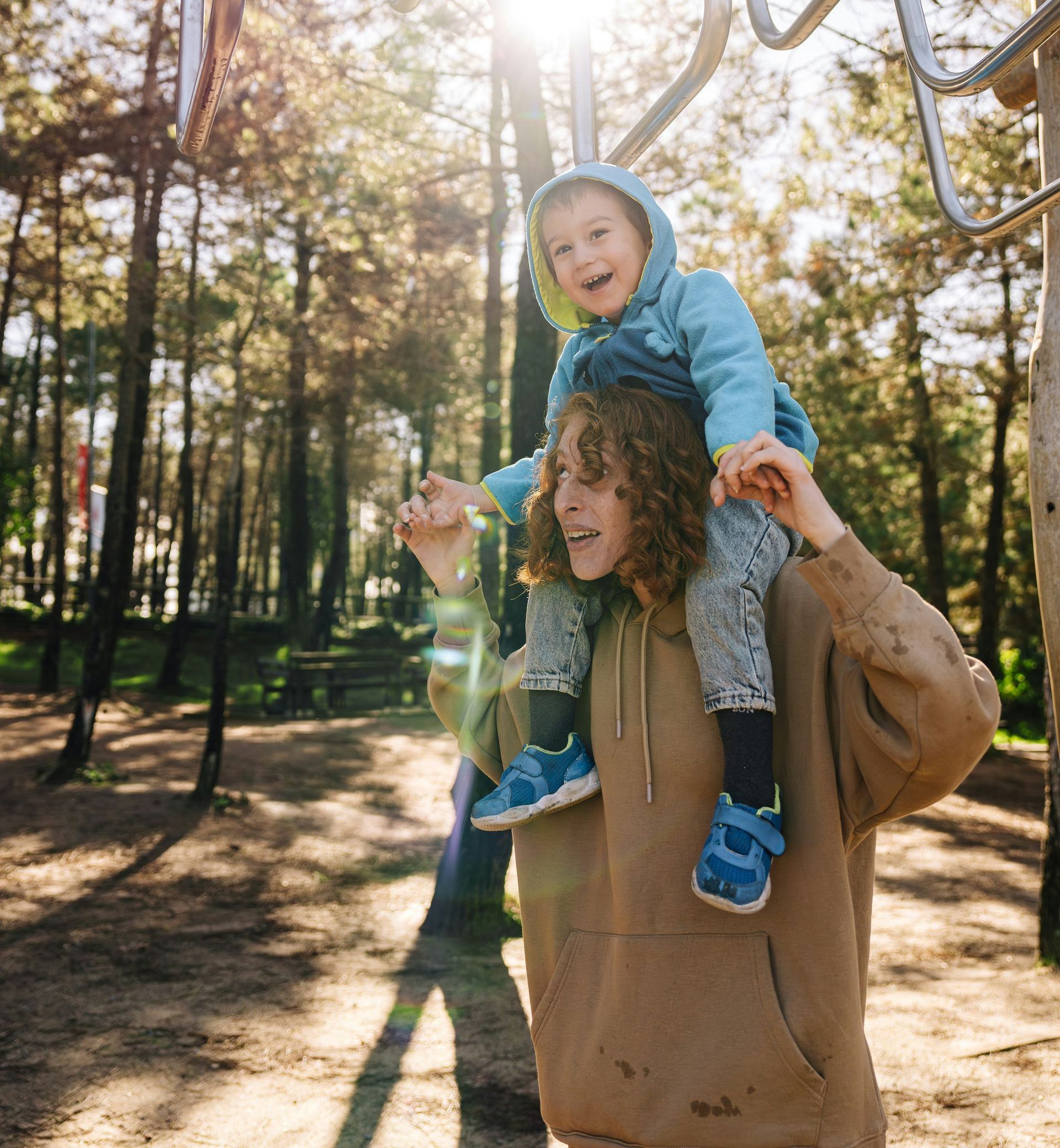 Adult carrying a smiling child on shoulders in a sunlit park with tall trees and long shadows