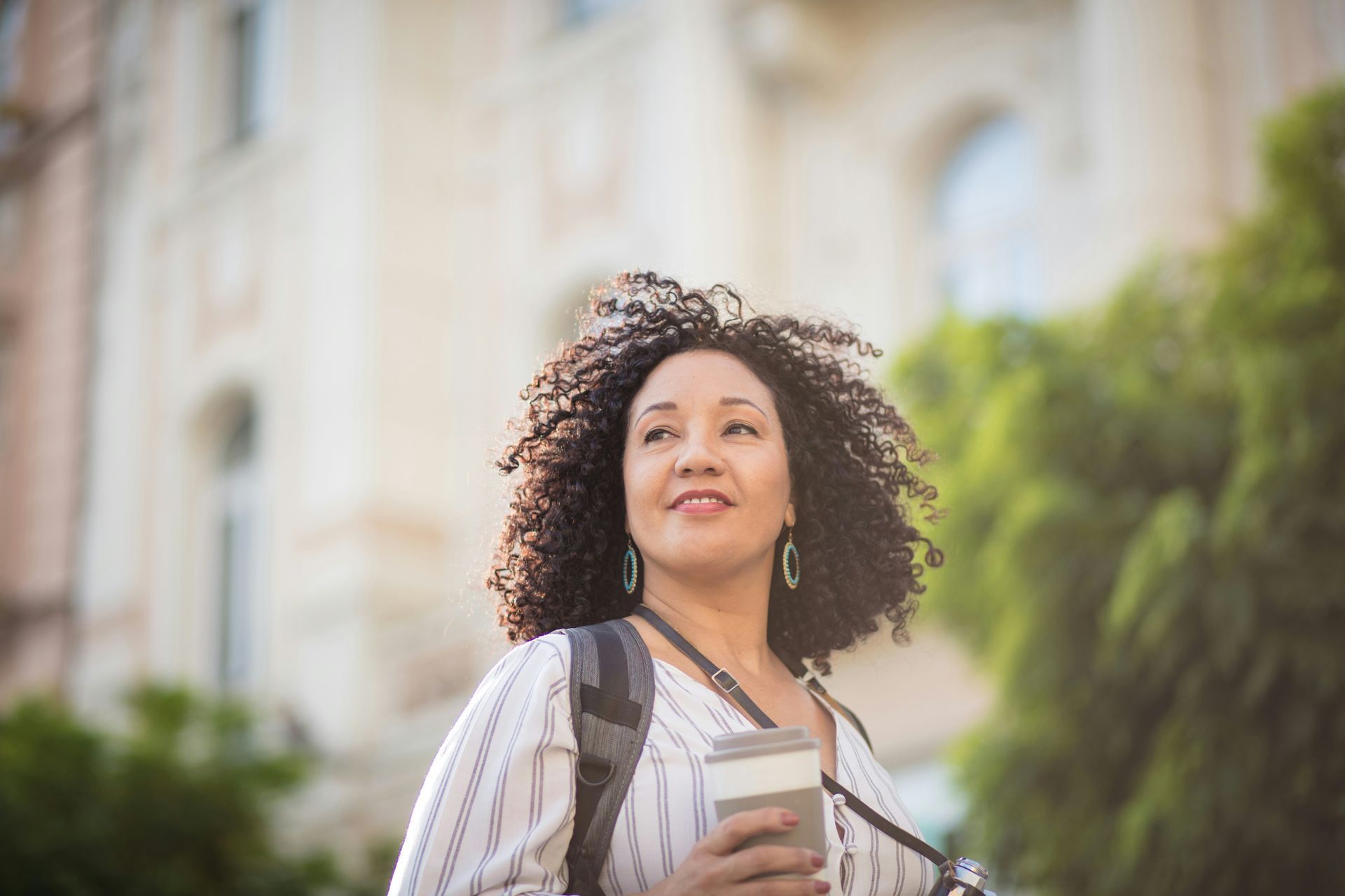 Woman holding a coffee cup outdoors with a backpack, standing in front of a blurred building and trees
