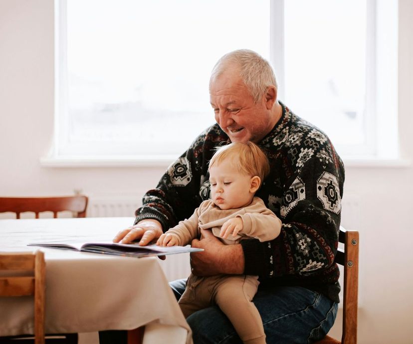 Older man smiling at a toddler on his lap while reading at a table in a bright room