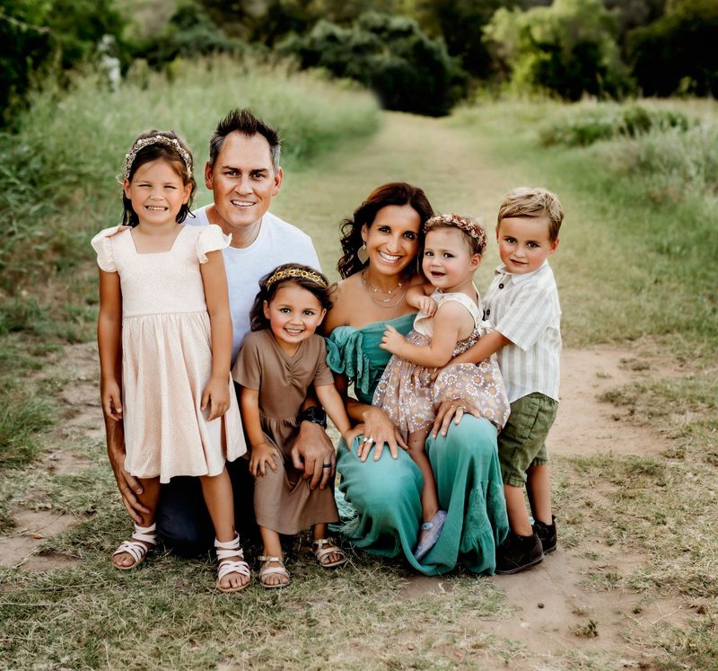 Family of five posing outdoors on a grassy path, smiling and dressed in light summer clothes.