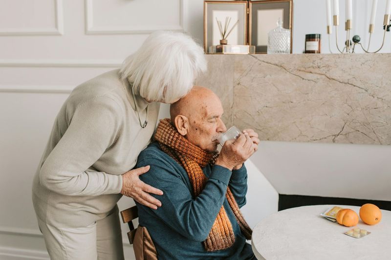 Two older people in a kitchen, one helping the other drink from a cup at a table with oranges.