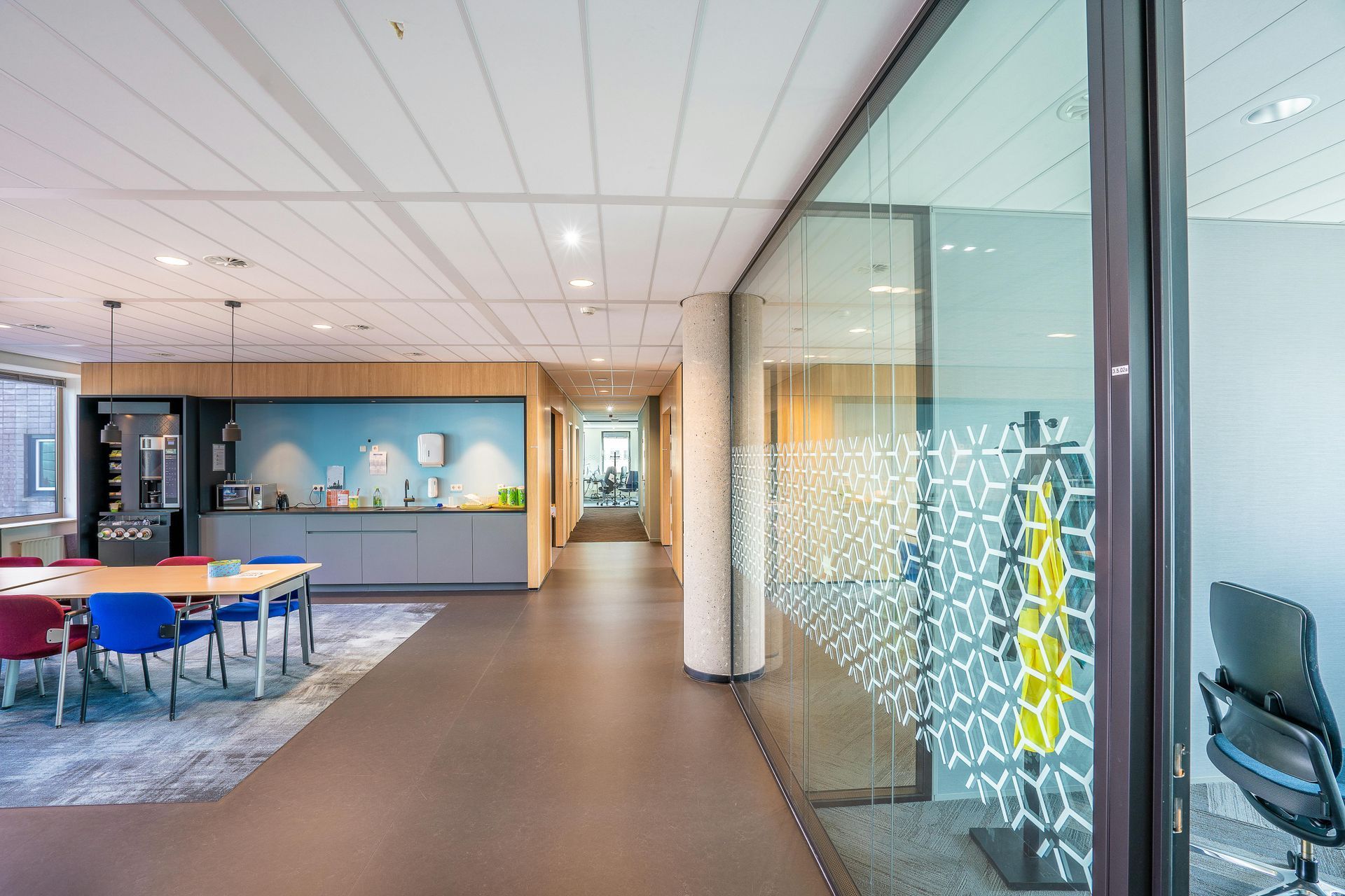 Office hallway with a seating area, pantry, and glass-walled conference room. The hallway is dark, with light-colored walls.