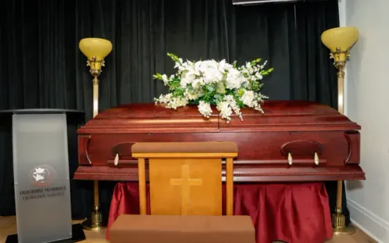 Closed coffin on a stage with white flowers, flanked by lamps and a podium draped in red cloth.