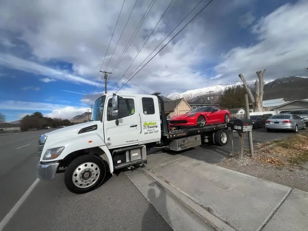White tow truck carrying a red car, parked by a curb near a residential street under cloudy skies
