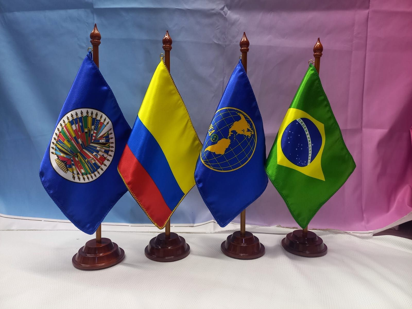 Four tabletop flags: OAS, Colombia, OAS, and Brazil on wooden bases against a pink backdrop