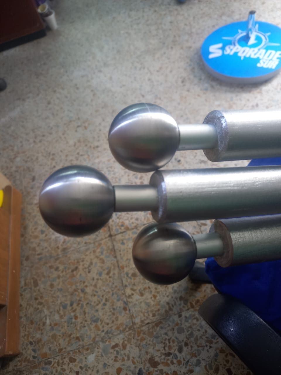 Three metal bars with silver ball ends on a speckled floor, next to a blue base and a round blue sign.