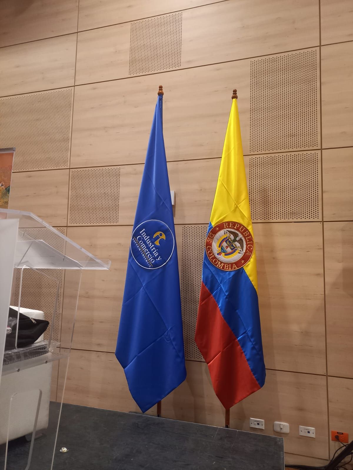 Two standing flags, blue and Colombia’s tricolor, beside a clear podium in a conference room.